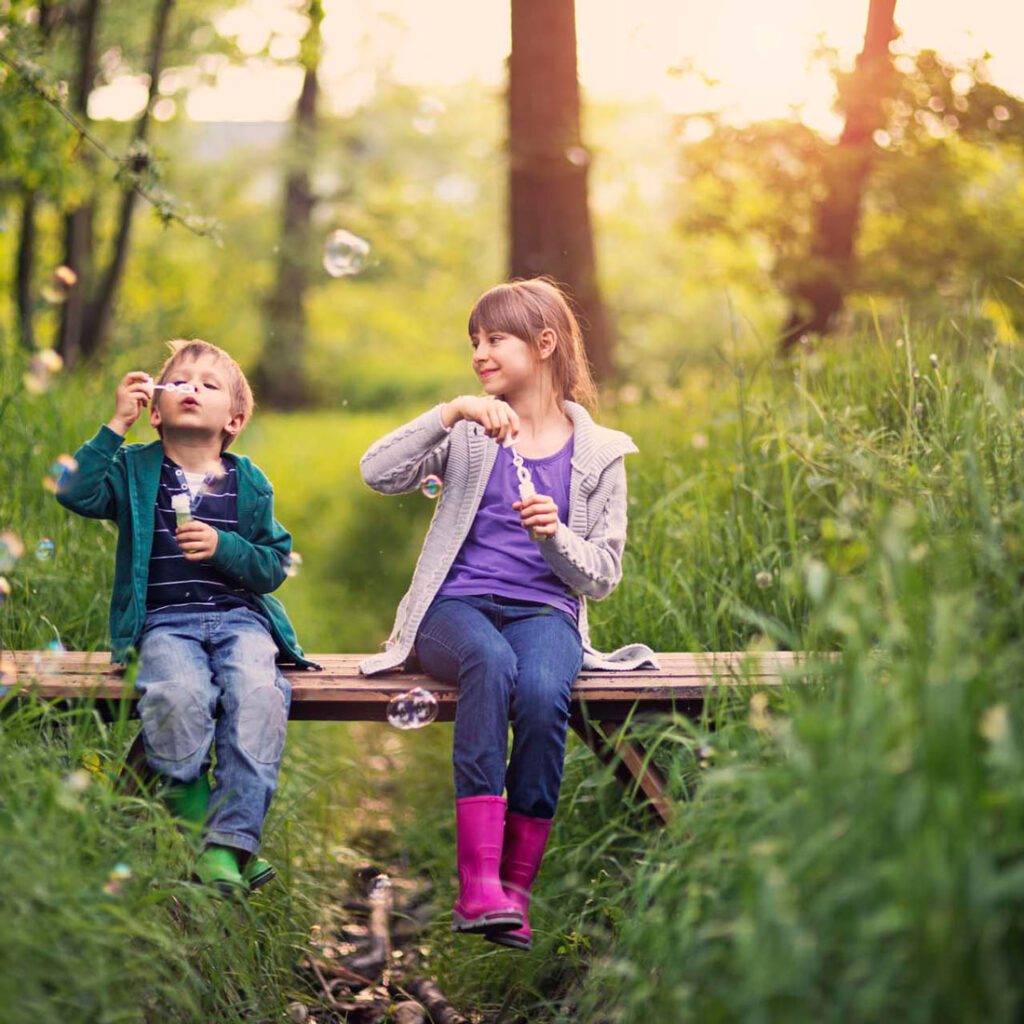 A boy and a girl blowing bubbles on a wooden bench in the woods, a playful scene of sibling connection in adoption