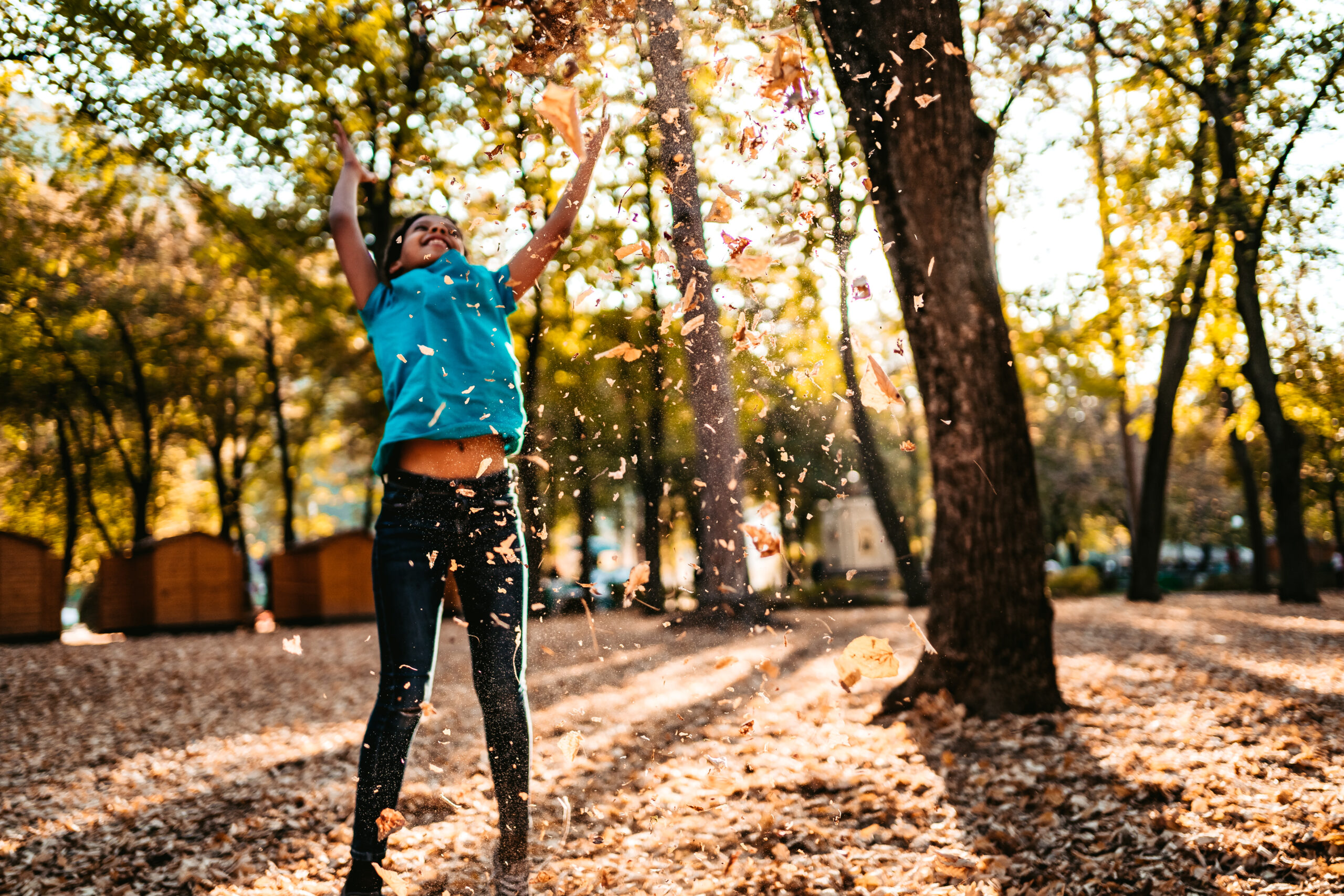 Child joyfully tossing autumn leaves into the air in a sunlit park, surrounded by tall trees and golden light. The child is wearing a blue top and dark trousers, with a big smile and arms raised high.